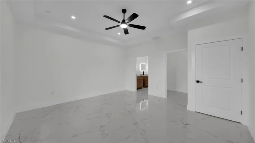 Unfurnished bedroom with a tray ceiling, connected bathroom, light marble finish flooring, ceiling fan, and recessed lighting