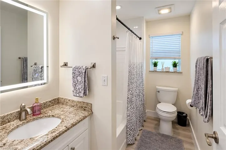 Bathroom featuring an illuminated mirror, a vanity with a stone-finish countertop, a built-in tub with a shower, a window with blinds, and wood-finish flooring