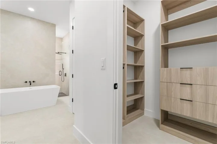 Integrated bath and closet featuring a freestanding tub, expansive walk-in shower with glass enclosure, built-in open shelving, and wood-finish cabinetry with matte black hardware
