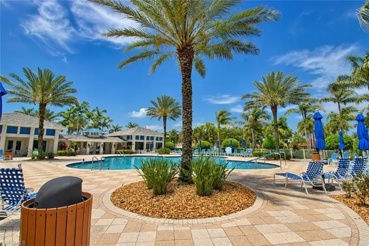 Resort-style pool area featuring a large swimming pool, surrounding paver patio, lush landscaping with palm trees, and multiple lounge chairs with blue umbrellas