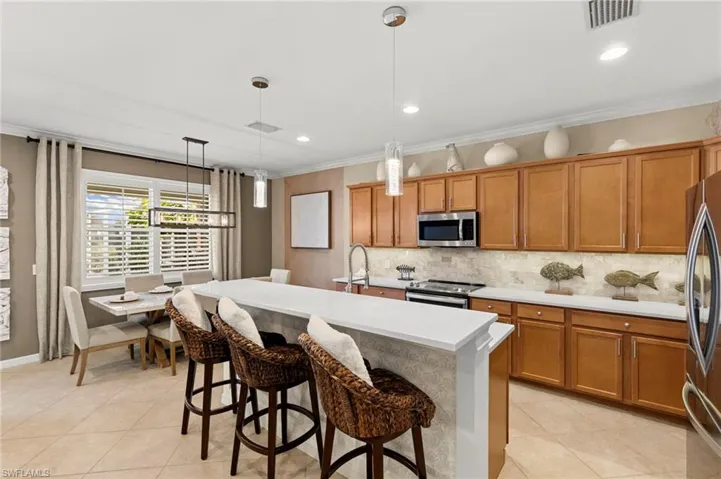 Kitchen featuring brown cabinetry, hanging light fixtures, stainless steel appliances, a center island with sink, and crown molding