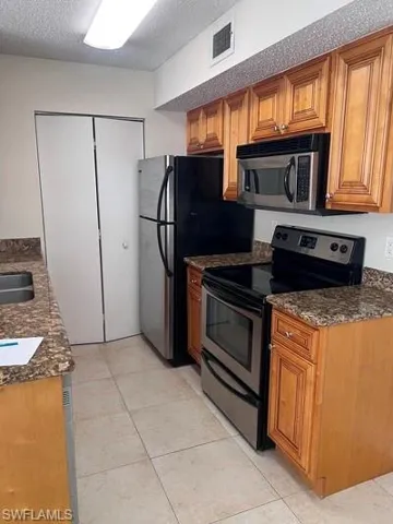 Kitchen featuring stainless steel appliances, dark stone countertops, wood finish cabinets, a textured ceiling, and light tile patterned floors