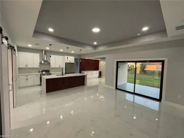 Kitchen featuring a barn door, a tray ceiling, light marble finish flooring, recessed lighting, and a kitchen island with sink