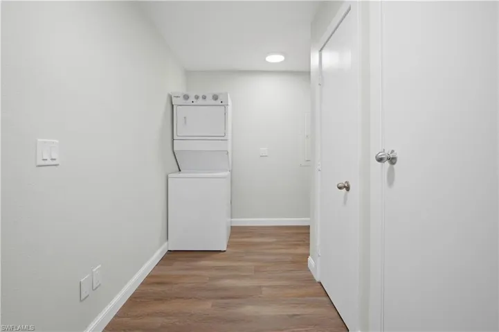 Laundry area featuring wood-finish flooring, a stacked washer and dryer unit, white baseboards, and a ceiling-mounted light fixture