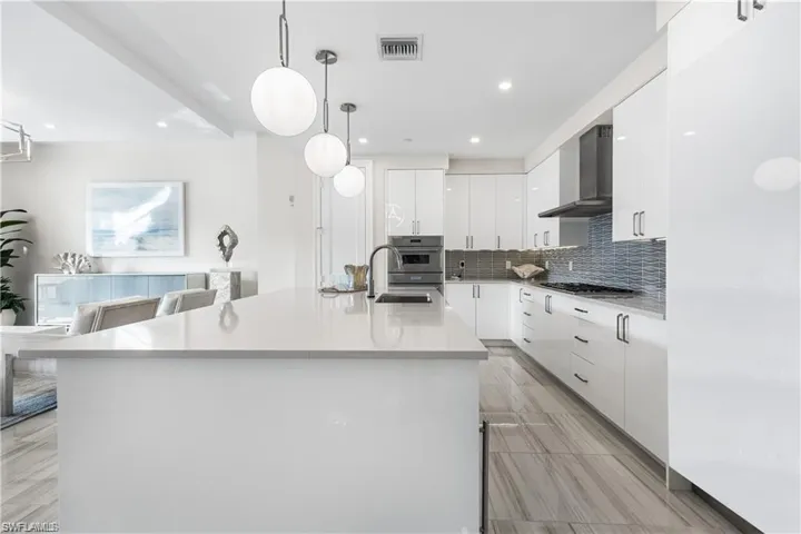 Kitchen featuring sink, wall chimney exhaust hood, hanging light fixtures, a center island with sink, and white cabinets