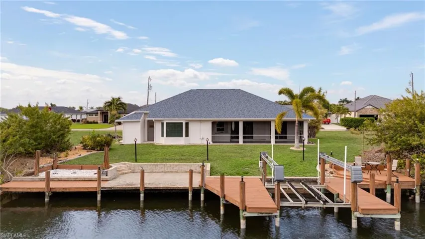 Rear view of property featuring a water view, boat lift, a lawn, a sunroom, and roof with shingles