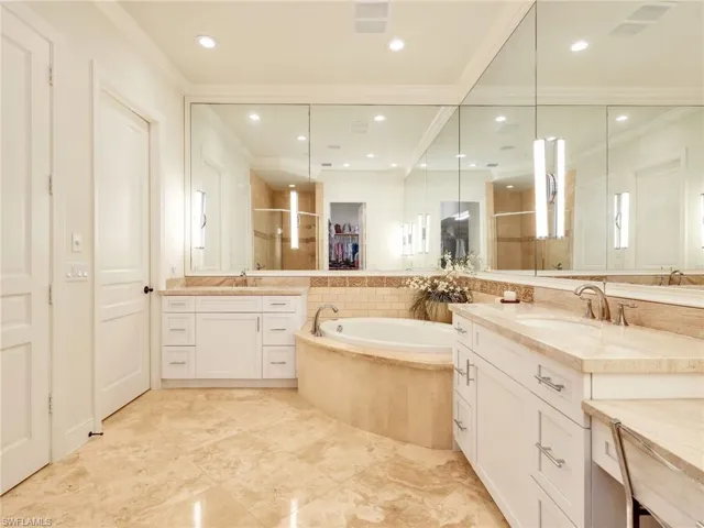 Owner's Suite Bathroom featuring marble flooring, a large corner jacuzzi tub, dual white vanity cabinets with stone countertops, and extensive mirrored wall panels.