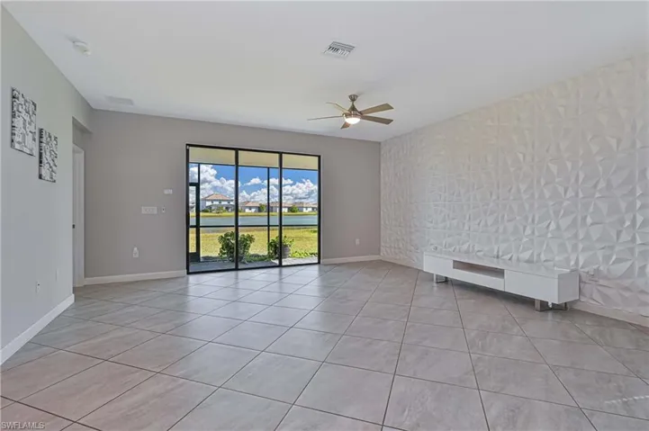 Large living area featuring neutral tile flooring, a textured geometric accent wall, ceiling fan, and floor-to-ceiling glass sliding doors with water scenery
