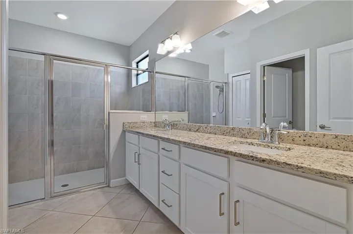 Dual-sink vanity with stone-finish countertops, white cabinetry, full-width vanity mirror, walk-in shower with tile surround, and tile flooring