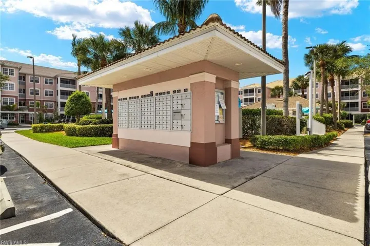Community mailbox structure featuring multiple mail receptacles and a tiled roof, surrounded by manicured landscaping, paved walkways, and palm trees