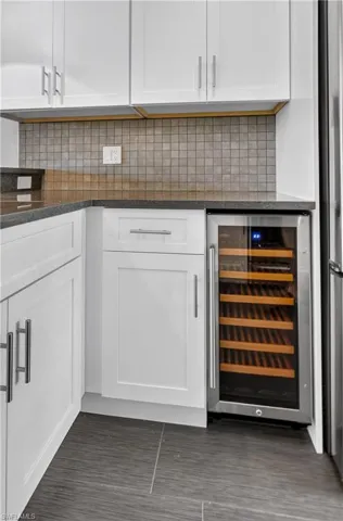 Stainless steel beverage cooler built into white shaker cabinetry with dark stone countertops, mosaic tile backsplash, and large format floor tiling