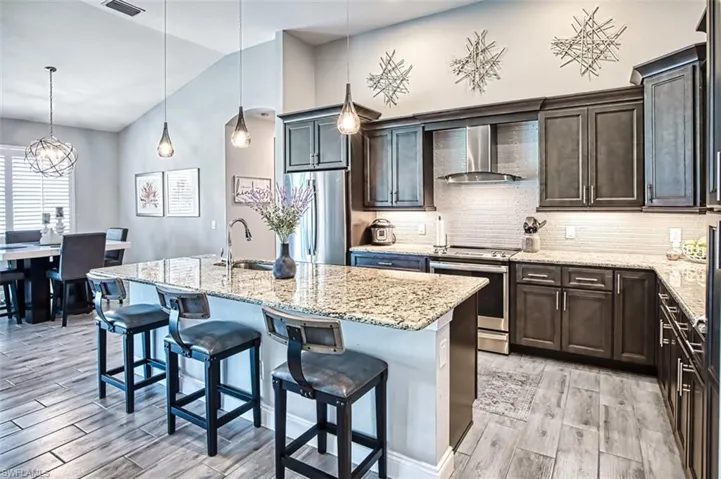 Kitchen with a center island with sink, appliances with stainless steel finishes, wall chimney exhaust hood, decorative light fixtures, and vaulted ceiling