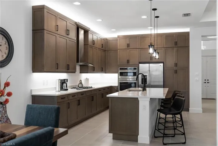 Kitchen with stainless steel appliances, hanging light fixtures, wall chimney exhaust hood, light tile patterned floors, and a kitchen island with sink