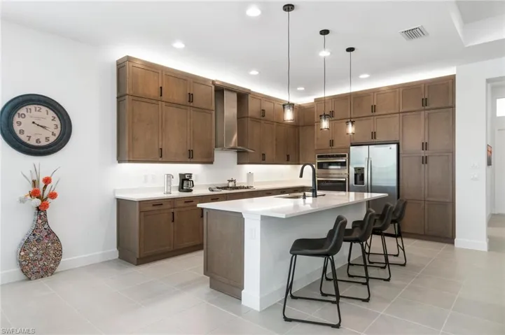 Kitchen featuring wall chimney range hood, an island with sink, stainless steel appliances, decorative light fixtures, and sink