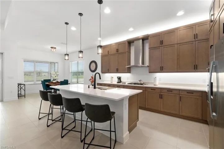 Kitchen with wall chimney range hood, hanging light fixtures, sink, a kitchen breakfast bar, and a kitchen island with sink