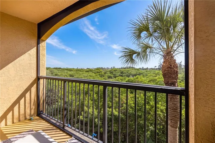 Balcony with stucco walls, arched opening, and black metal railings overlooking lush greenery and a palm tree