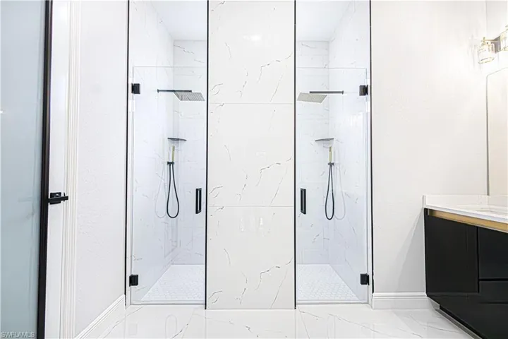 Modern bathroom featuring two glass-enclosed showers with rainfall showerheads and handheld sprayers, set against a backdrop of white marble-look tile walls and white hexagonal floor tiles