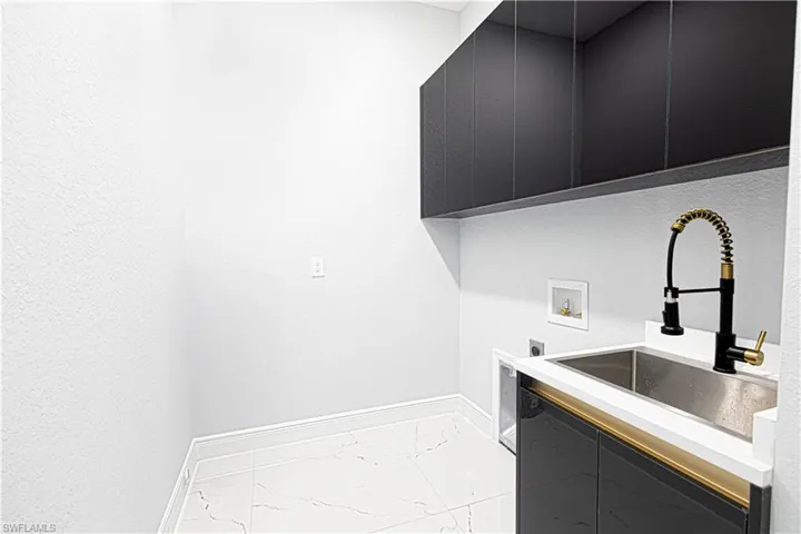 Utility area featuring a stainless steel sink with a modern black and gold faucet, dark cabinetry, and white tile flooring