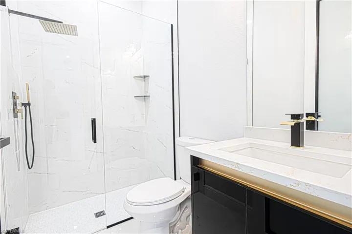 Bathroom featuring a glass-enclosed shower with a rainfall showerhead, a toilet, and a vanity with a white countertop and a contemporary black and gold faucet
