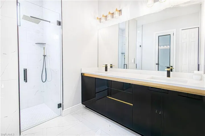 Bathroom featuring a glass-enclosed shower with a rainfall showerhead, a dual-sink vanity with a gold and black finish, and a large mirror