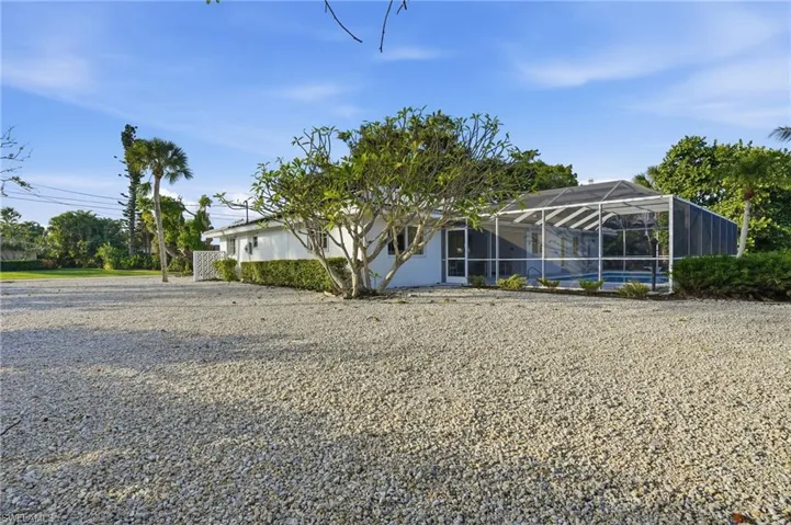 White stucco exterior with a screened-in pool enclosure, surrounded by mature tropical landscaping and a gravel yard