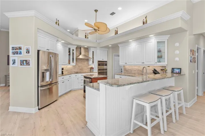 Kitchen with appliances with stainless steel finishes, light stone counters, white cabinets, kitchen peninsula, and wall chimney range hood