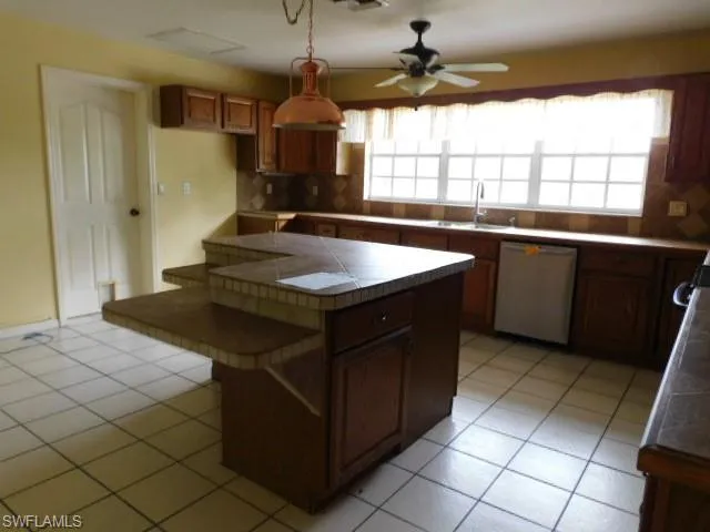 Kitchen featuring tile countertops, backsplash, brown cabinetry, and dishwashing machine