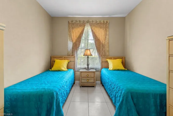 Room featuring neutral wall tones, large window with patterned drapes, tile flooring, and a two-drawer nightstand with a lamp