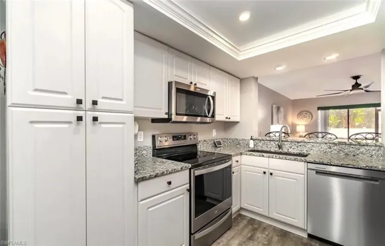 Kitchen with appliances with stainless steel finishes, light stone counters, white cabinets, a tray ceiling, and dark wood-type flooring