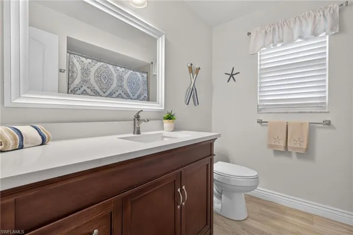Bathroom featuring a wood-finish vanity with a white countertop and integrated sink, a large framed mirror, a toilet, a window with horizontal blinds, and tile flooring