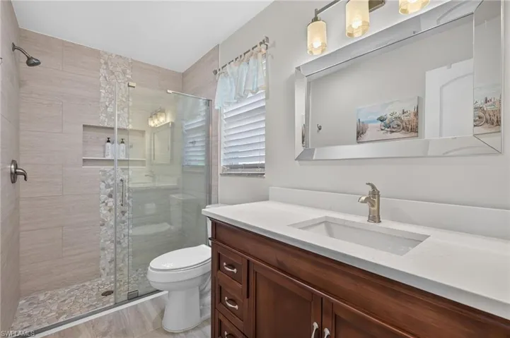 Bathroom featuring a frameless glass shower enclosure, stone tile shower surround with pebble floor, built-in shower niche, vanity with an integrated sink, and a rectangular mirrored vanity fixture
