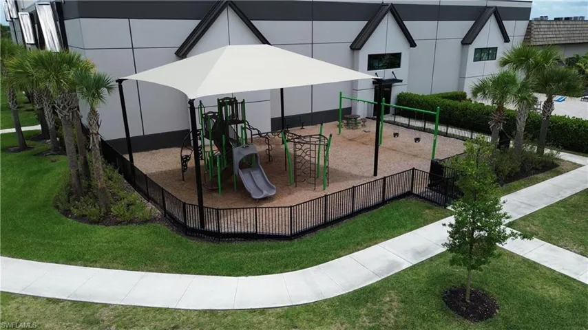 Fenced recreational area featuring a playground with a slide and swings, shaded by a large canopy structure