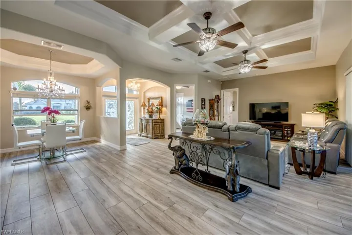 Living room with light wood-type flooring, coffered ceiling, ceiling fan with notable chandelier, crown molding, and beamed ceiling