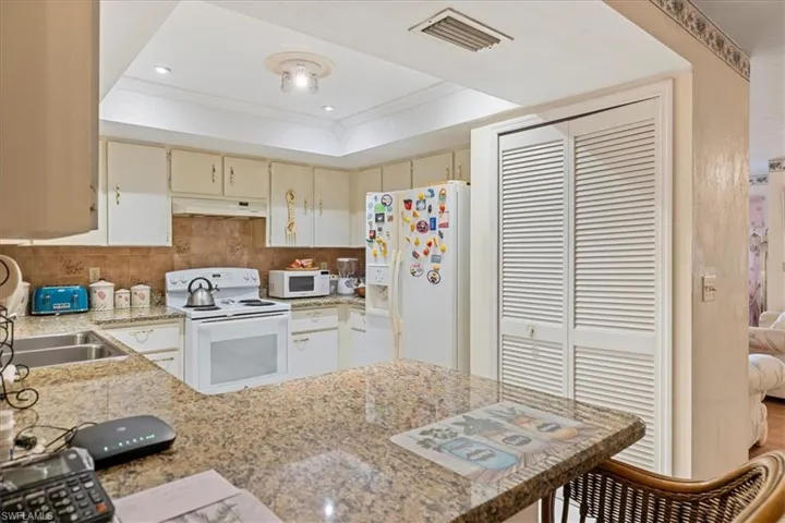 Kitchen featuring cream cabinetry, white appliances, kitchen peninsula, light stone countertops, and a tray ceiling