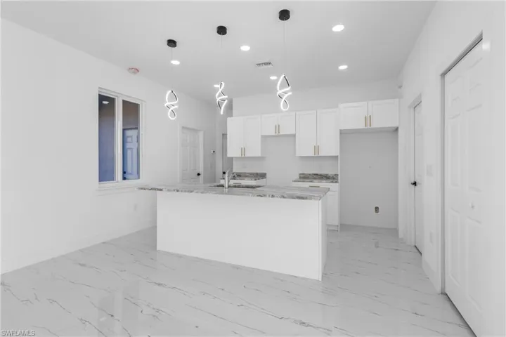 Kitchen with white cabinetry, light stone countertops, hanging light fixtures, a center island with sink, and light marble finish floors