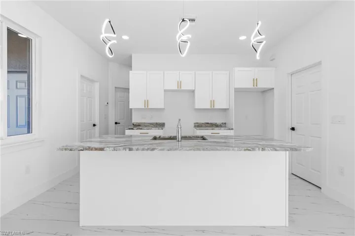 Kitchen with white cabinetry, light stone counters, decorative light fixtures, and a center island with sink