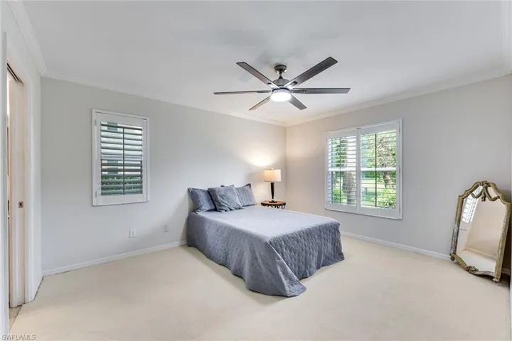 Light-filled room featuring neutral wall tones, crown molding, a ceiling fan with integrated lighting, two windows with plantation shutters, and light-toned carpet flooring