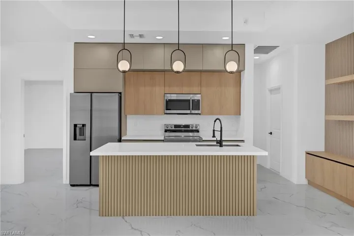 Two tone kitchen with modern cabinets, pendant lighting, stainless steel appliances, a kitchen island with sink, and light stone countertops