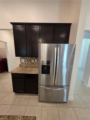 Stainless steel French door refrigerator, dark wood-finish cabinetry, granite countertop, mosaic tile backsplash, and tile flooring