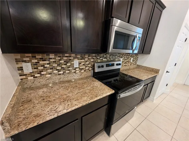 Dark wood-finish cabinetry, granite countertops, mosaic tile backsplash, stainless steel microwave, and a black electric range