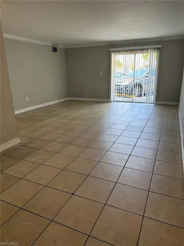 Spacious room featuring neutral tone walls, tile flooring, white baseboards, crown molding, and a sliding glass door with vertical blinds