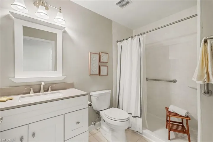 White vanity cabinet with an integrated rectangular sink and stone-finish countertop, three-light vanity fixture, framed wall mirror, and walk-in shower featuring a metal support bar