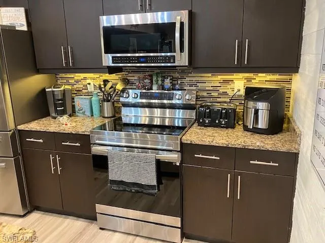 Kitchen with stainless steel appliances, dark brown cabinetry, backsplash, light stone counters, and light wood-style floors