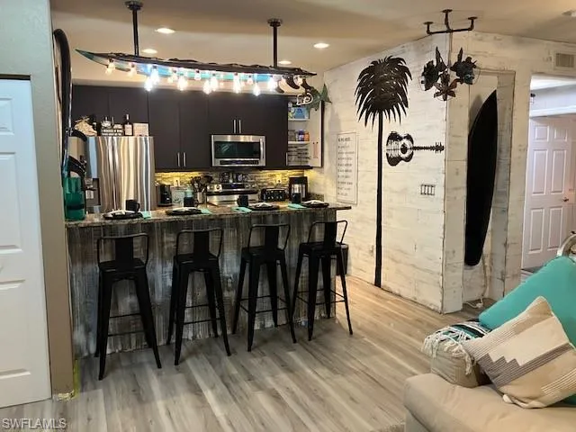 Kitchen with a kitchen bar, light wood-style flooring, stainless steel appliances, open shelves, and hanging light fixtures