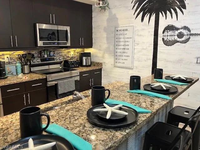 Kitchen featuring appliances with stainless steel finishes, dark cabinetry, light stone countertops, wooden walls, and tasteful backsplash
