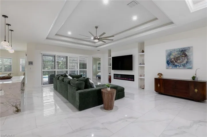 Living room with a raised ceiling, ornamental molding, built in shelves, light marble finish floors, and a ceiling fan