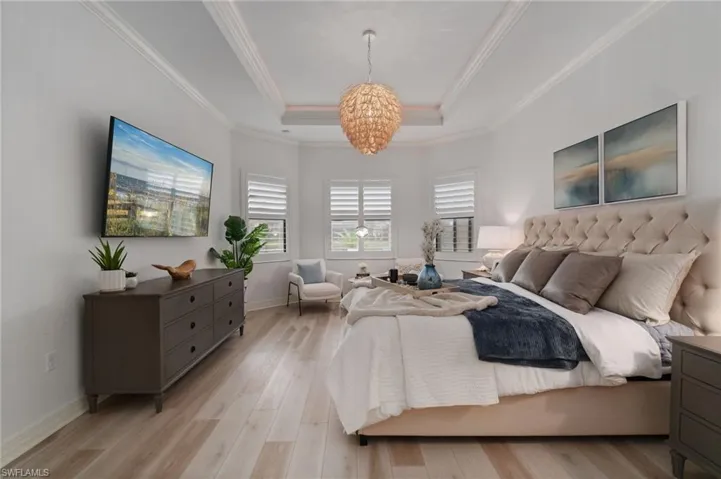 Bedroom with a raised tray ceiling accent, a coastal modern statement chandelier, favorable light wood-style floors, and decorative crown  molding.
