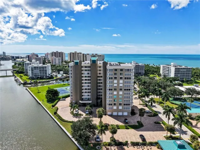 High-rise residential exterior featuring waterfront positioning, expansive paved parking areas, surrounding palm trees, and manicured green spaces