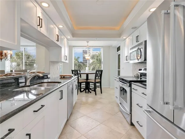 White cabinetry featuring matte black hardware, dark stone countertops, diamond-pattern tile backsplash, stainless steel dishwasher, and tray ceiling lighting