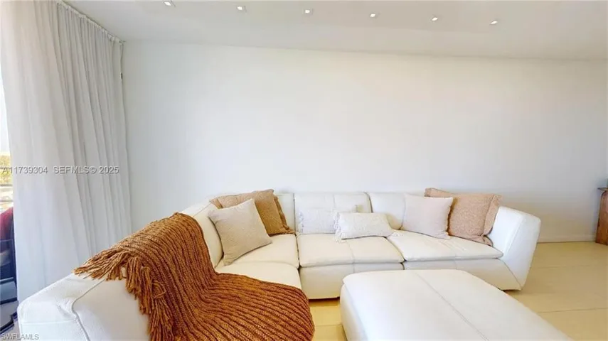 Recessed ceiling lighting illuminates a spacious room featuring a neutral color palette, light-toned flooring, and a large window with sheer drapes